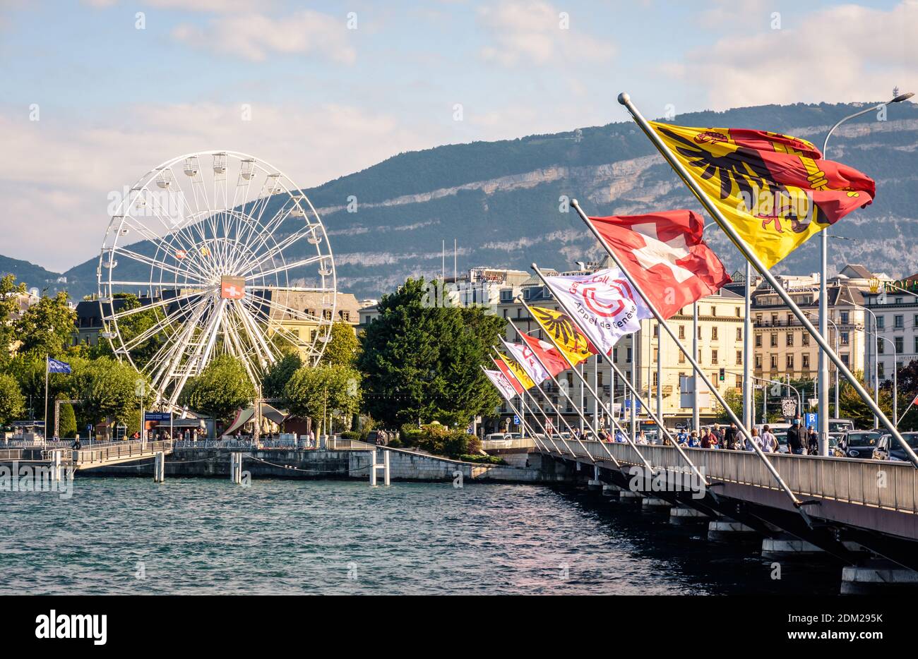 Genfer Flaggen und Schweizer Flaggen, die auf der Mont Blanc-Brücke auf dem Genfersee mit dem Riesenrad in der Ferne fliegen. Stockfoto