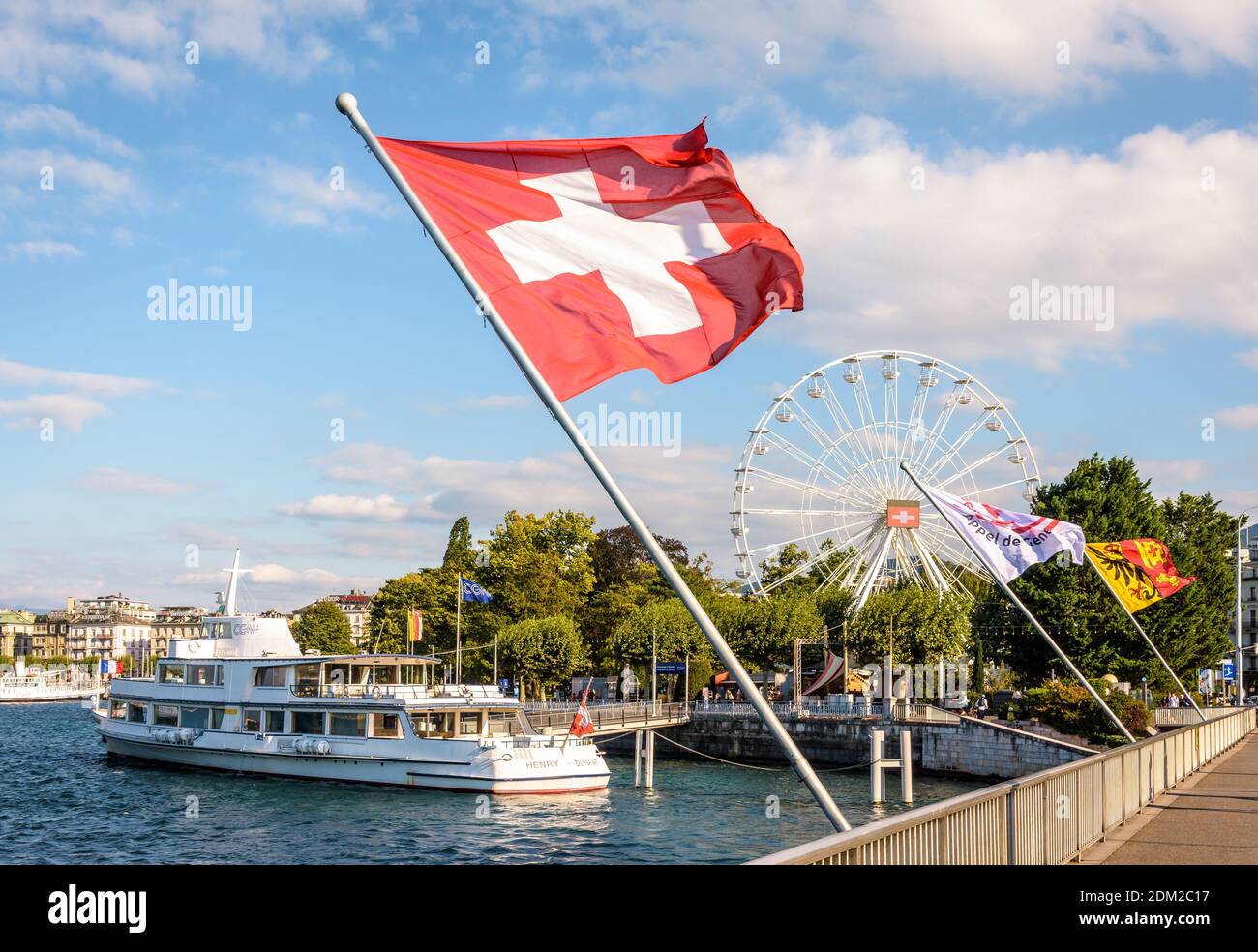 Die Schweizer Flagge fliegt im Wind auf dem Genfersee mit einem Ausflugsboot, das am Fuß des Riesenrads festgemacht ist. Stockfoto
