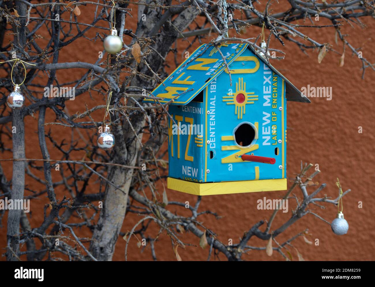 In Santa Fe, New Mexico, hängt ein Vogelhaus, das aus einem alten Nummernschild gefertigt wurde, in einem Baum, der mit Weihnachtsornamenten geschmückt ist. Stockfoto