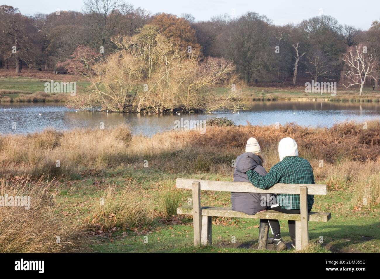 Ein junges Paar, das auf einer Parkbank sitzt und sich warm einwickelt, in der Nähe von Pen Ponds im Richmond Park, London, Großbritannien Stockfoto