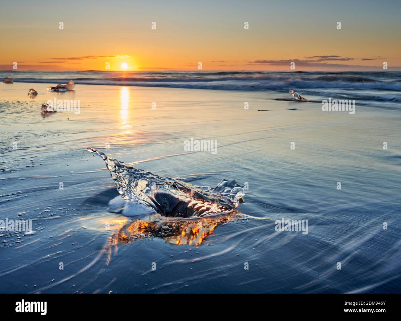 Klares Stück Gusseis am nassen Strand mit Meer Wellen im Sonnenuntergang auf dem Hintergrund Stockfoto