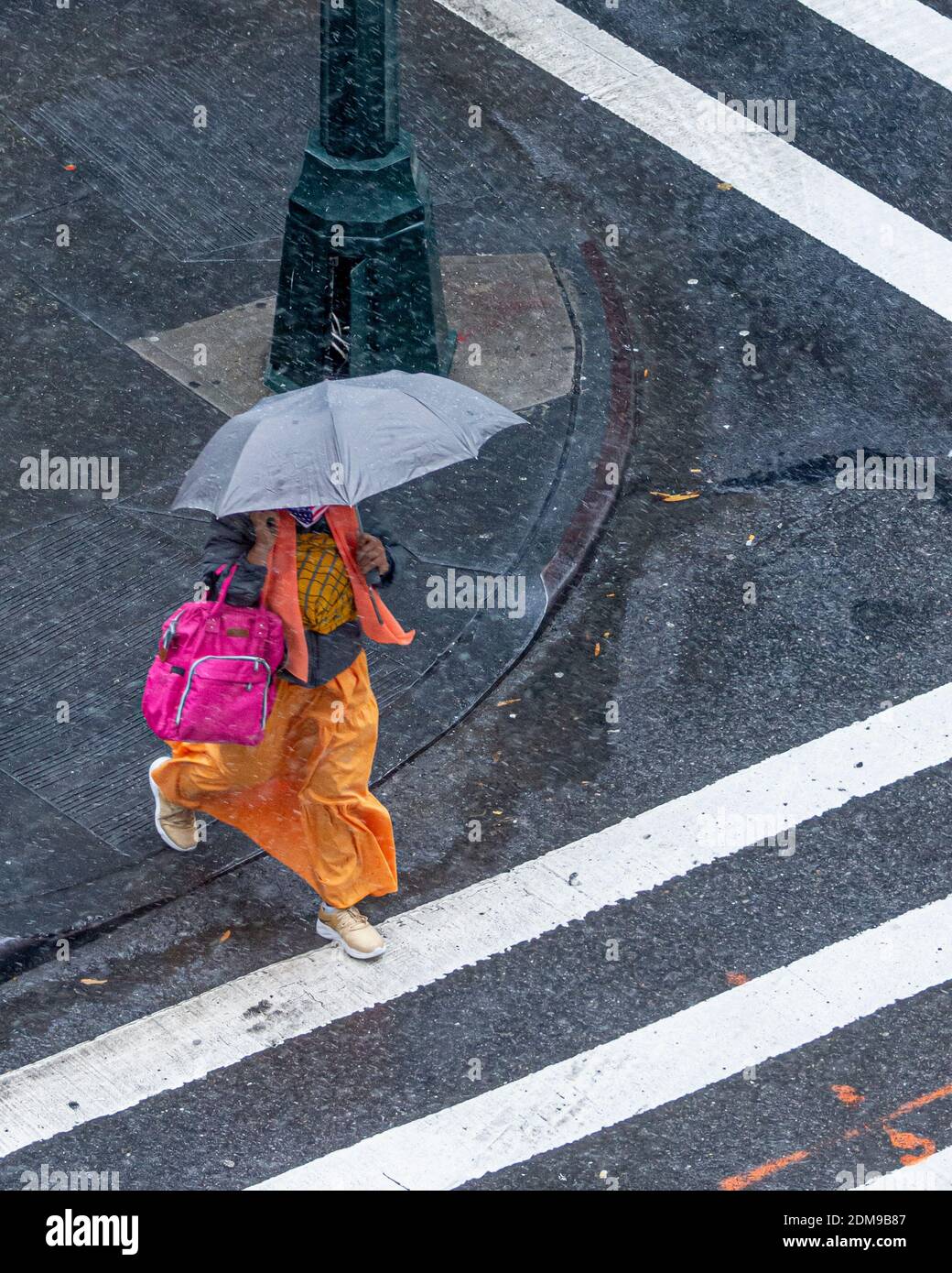 Blick von oben auf Frau in bunten Kleid mit Regenschirm überqueren Stadtstraße an einem tristen Tag als helle Kleidung kontrastiert scharf mit dunklen Bürgersteig. Stockfoto