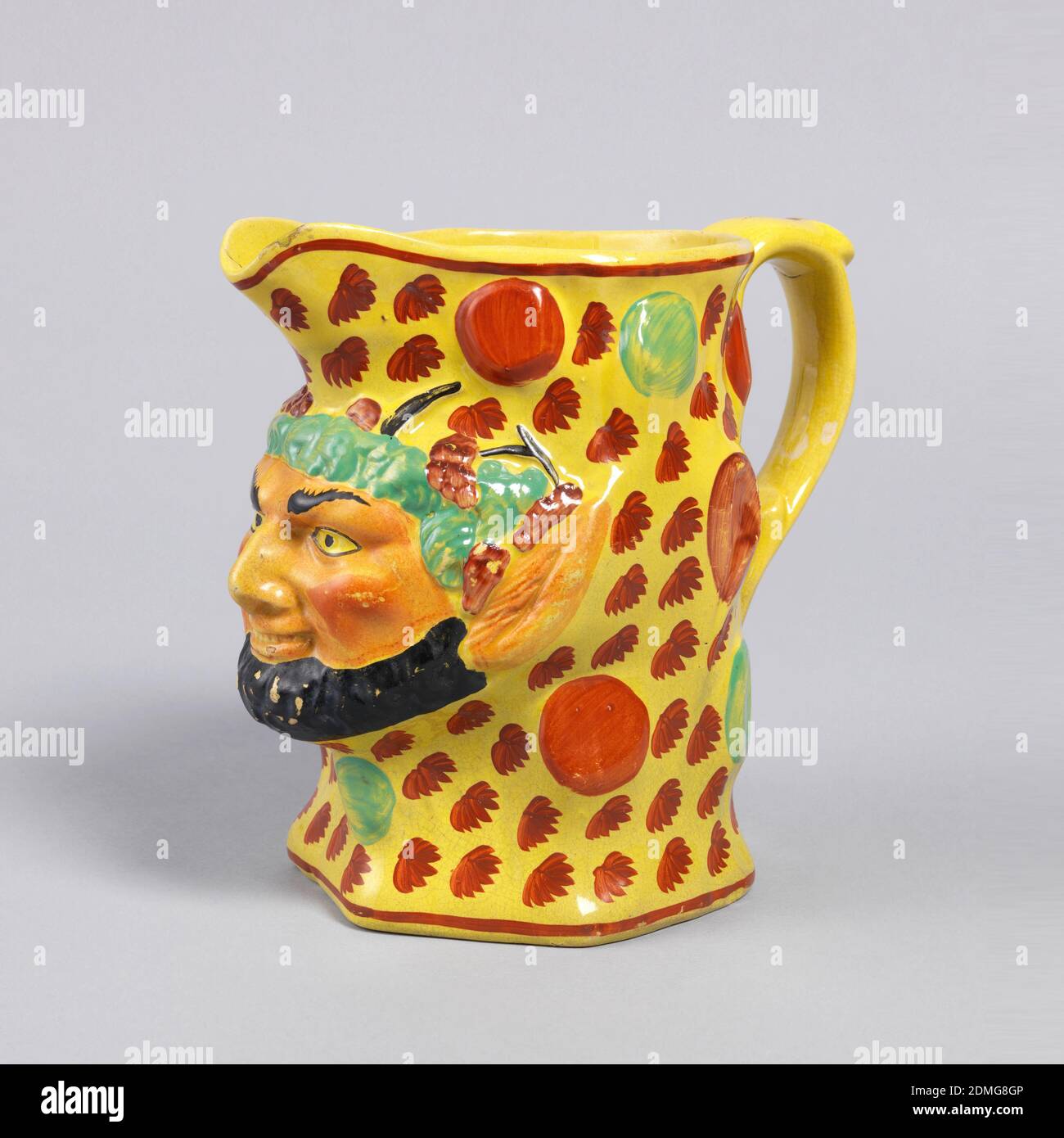 Krug, gelber Boden und bunt bemalte Dekoration auf geformtem Steinzeug, Satyr Maske mit schwarzem Bart mit spitzen Ohren und grünem Haar Projekte aus Krug. Der Rest der Kanne ist mit roten Blättern und Kreisen auf gelbem Boden verziert. Offener Auslauf; roter Rand um Rand und Sockel., England, 1820–60, Keramik, Dekorative Künste, Krug Stockfoto