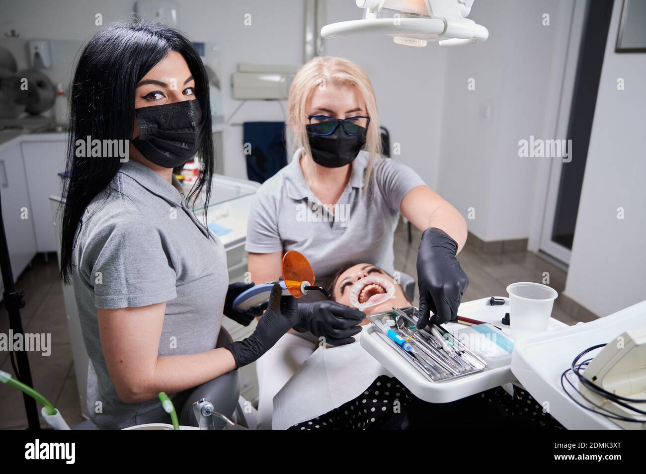 Schöne weibliche Assistentin in medizinische Maske Blick auf Kamera, während Arzt Befestigung Klammern an Patienten Zähne in der Zahnarztpraxis. Konzept der Zahnmedizin und kieferorthopädische Behandlung. Stockfoto