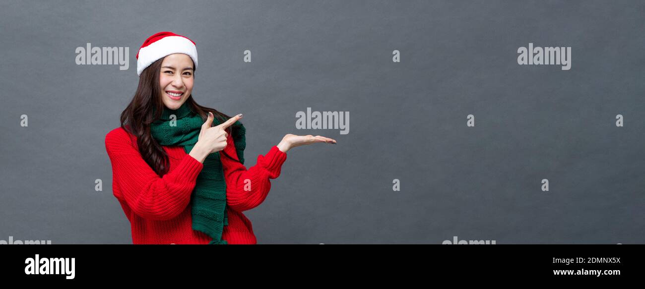 Studio Porträt einer fröhlichen asiatischen Frau trägt weihnachten Thema Kleidung zeigt Hand zu öffnen Handfläche mit leerem Kopieplatz Auf grauem Banner-Hintergrund Stockfoto