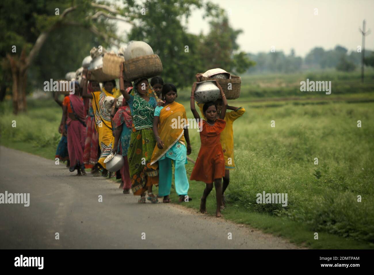 Eine Gruppe von Frauen und Kindern, die Bambuskörbe mit Metallgläsern auf dem Kopf tragen, während sie gemeinsam in einer Reihe entlang einer Straße laufen, die Bodh Gaya und Rajgir in Bihar, Indien verbindet. Stockfoto