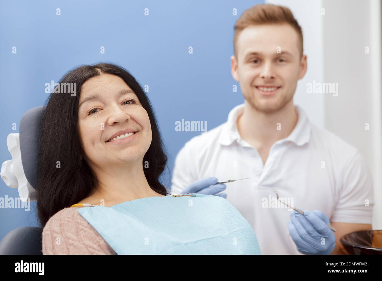 Entspannt beim Zahnarzt. Glückliche ältere Patientin sitzt in einem Zahnarztstuhl lächelnd zur Kamera ihr Zahnarzt lächelt auf dem Hintergrund hält seine Stockfoto