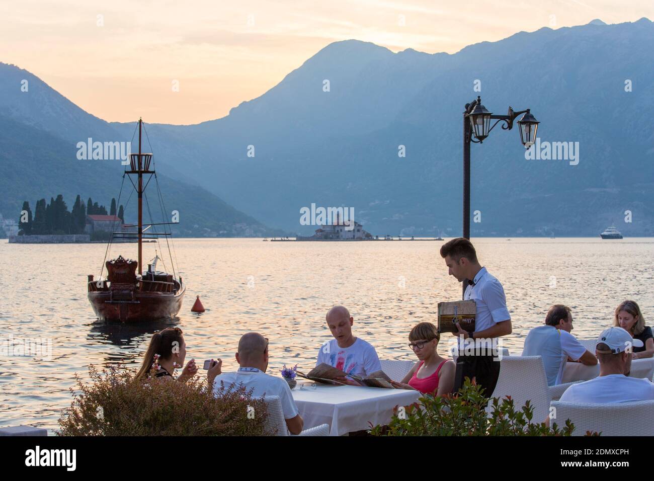 Perast, Kotor, Montenegro. Besucher, die Essen in einem Restaurant am Wasser mit Blick auf die Bucht von Kotor bestellen, Abenddämmerung. Stockfoto