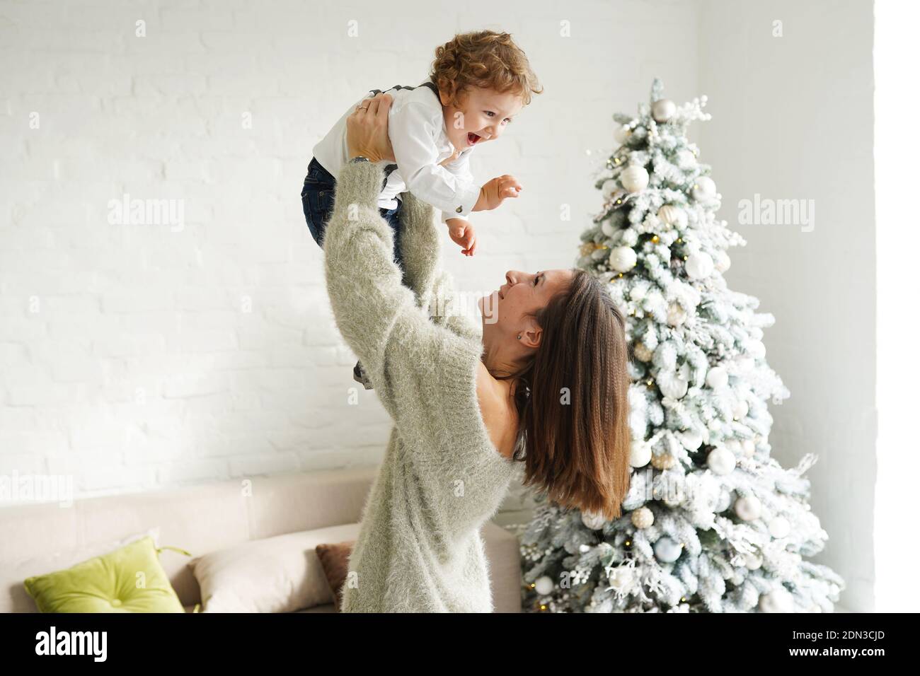 Ein süßer kleiner Junge lacht glücklich, während Mama hält Er über ihrem Kopf in der Nähe von weihnachtsbaum im Wohnzimmer Stockfoto