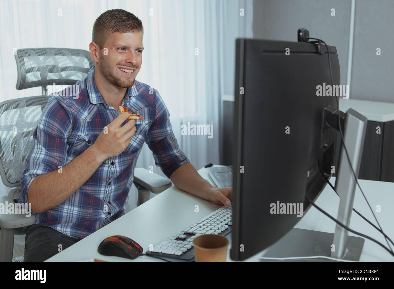 Handsome bearded IT industry worker enjoying working online, eating pizza in front of computer. Happy young man studying, eating pizza Stockfoto