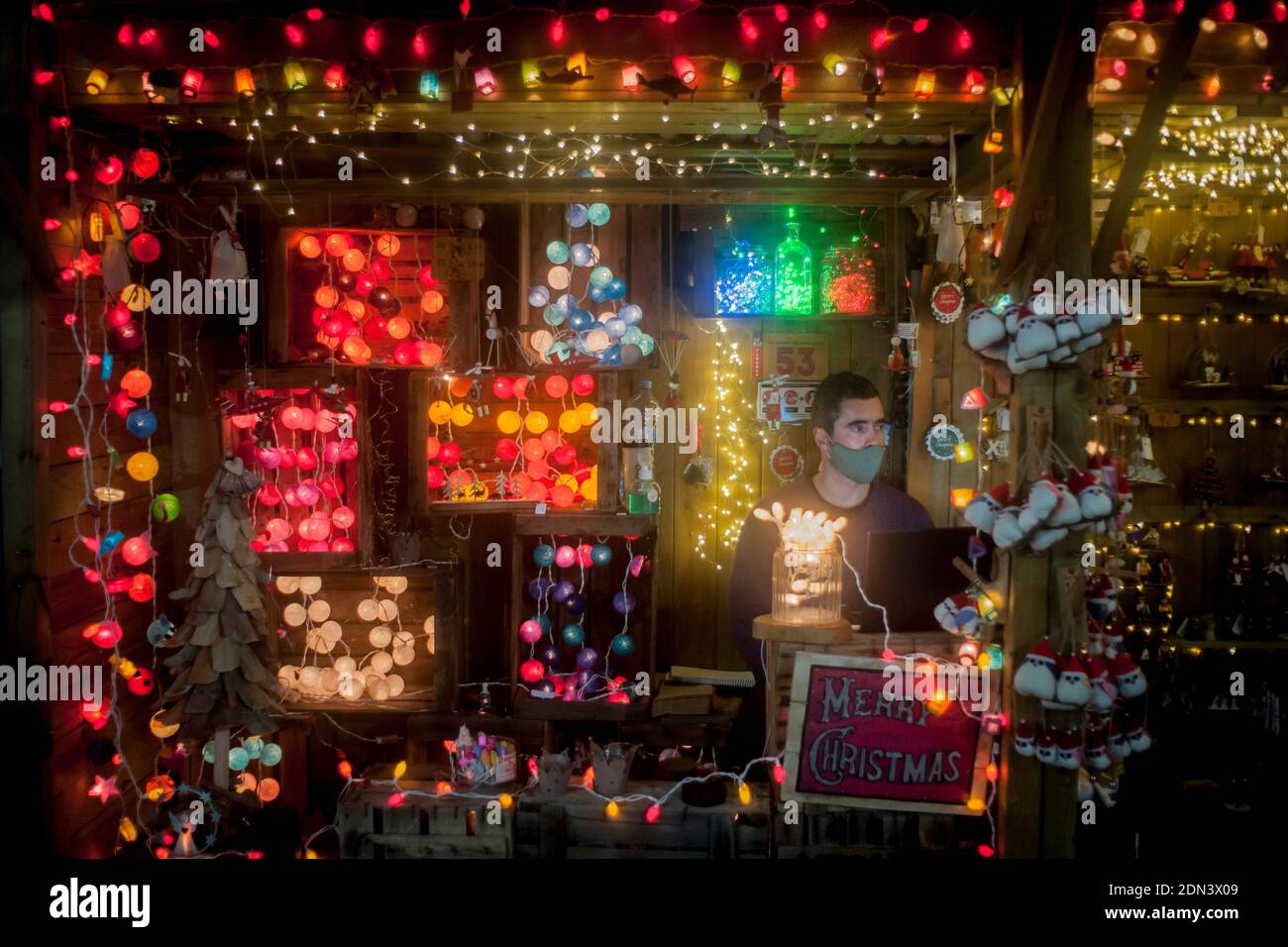 An einem dekorativen Lichterstand auf dem Weihnachtsmarkt von Santa Llucia in Barcelona wartet ein Verkäufer mit Gesichtsmaske auf Kunden. Stockfoto