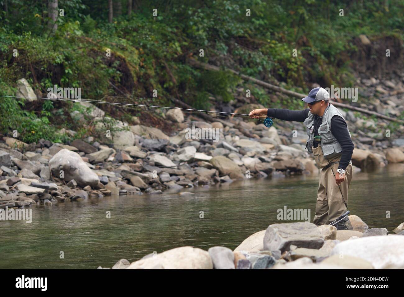 Fokussierter Fischer, der im Fluss steht und auf den Fang von Fischen auf Haken wartet. Mann im lässigen Outfit, der Freizeit für ruhige Aktivitäten in den Bergen verbringt. Stockfoto