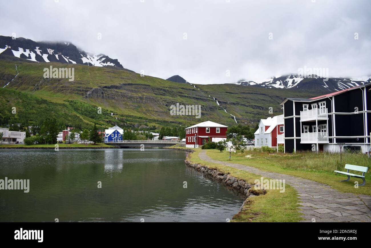 Blick über den See in Seydisfjordur auf die schneebedeckten Berge. Ostfjorde, Island. Ein Pfad führt um den Rand des Wassers. Stockfoto