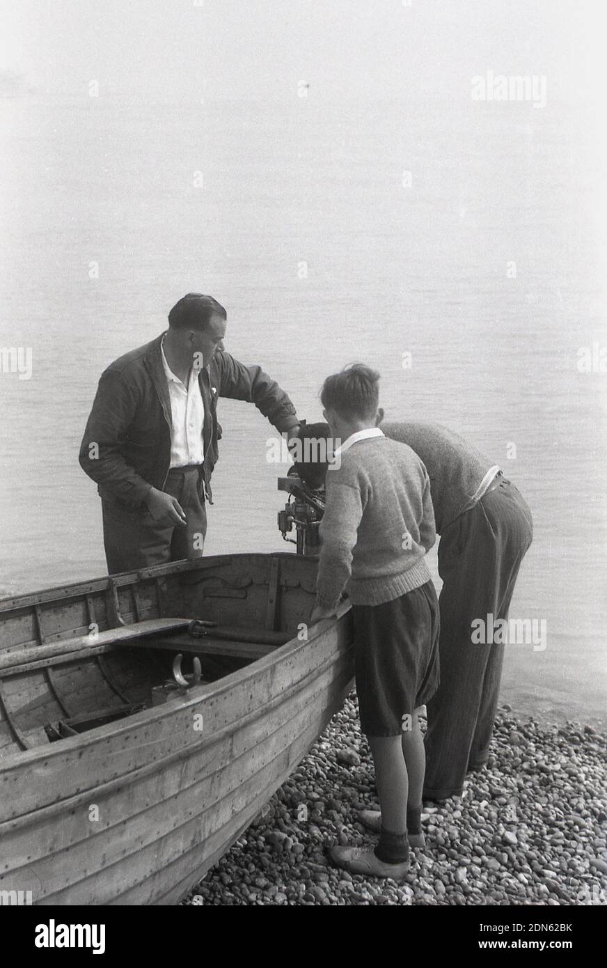1950er, historisch, zwei Männer und ein Jugendlicher stehen an einem hölzernen Ruderboot am Steinstrand, am Meeresrand bei Walmer, Deal, Kent, England, Großbritannien, Mit den Erwachsenen, um den Außenbordmotor zu überprüfen. Vielleicht brauchen sie es, denn die Wasserfläche vor ihnen ist der Ärmelkanal. Stockfoto