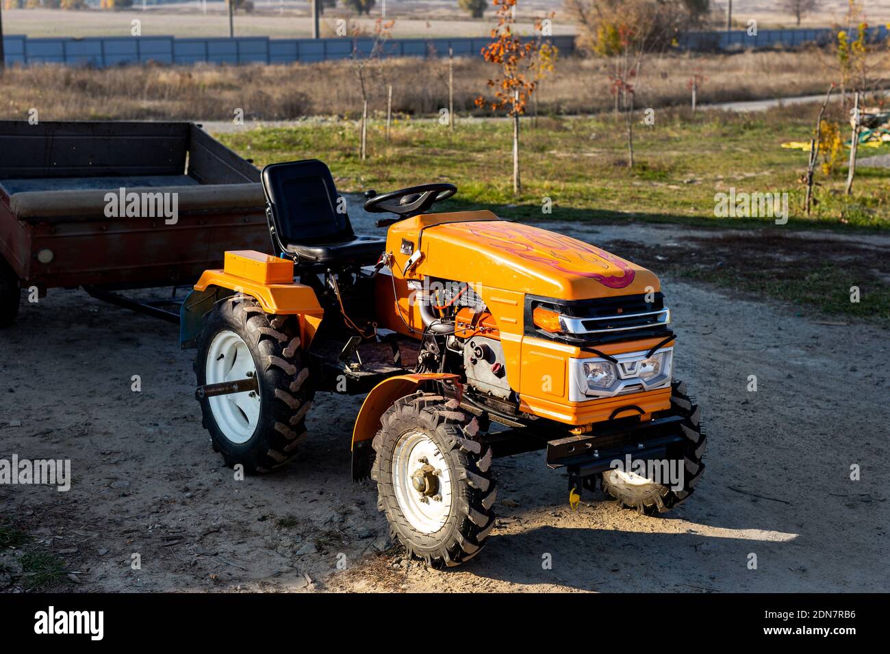Kleiner Mini-roter moderner orangefarbener neuer Traktor mit Anhänger, der in der Nähe des Hangargebäudes auf dem Land der Farm bei Sonnenuntergang oder Sonnenaufgang steht. Kleine landwirtschaftliche Betriebe Stockfoto