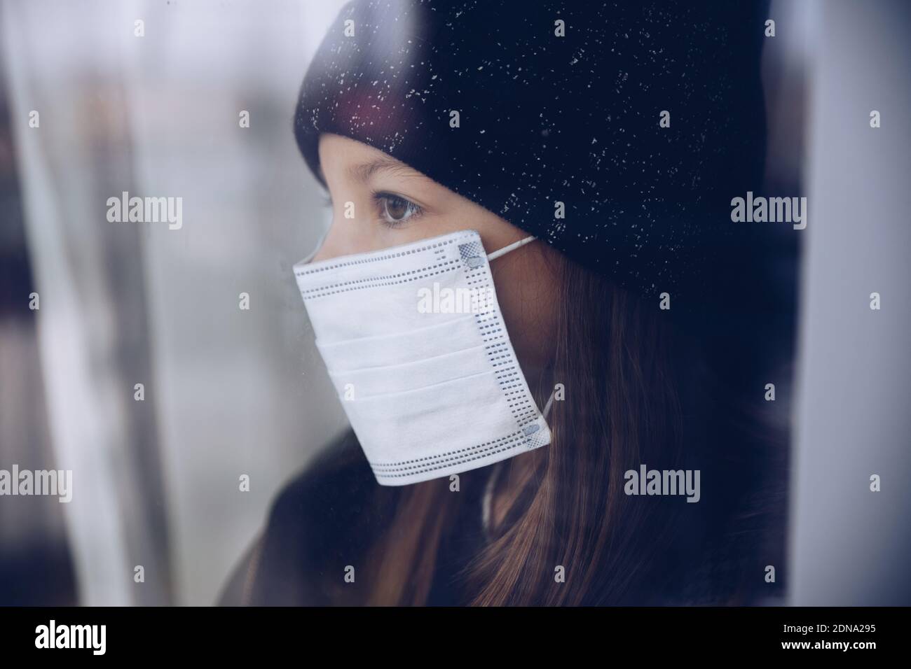 Ein schönes, trauriges Mädchen in einer Mütze und einer Schutzmaske wartet auf das Ende der Pandemie, um das Haus zu verlassen. Stockfoto
