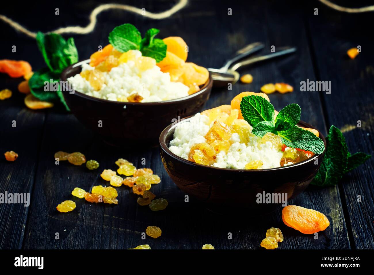 Hausgemachter Hüttenkäse mit getrockneten Aprikosen, Rosinen und Minze in Tonschüsseln, gesundes Frühstück, dunkler Hintergrund, selektiver Fokus Stockfoto