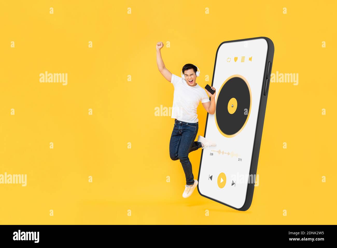 Glücklicher junger asiatischer Mann trägt Kopfhörer und hört Musik von Smartphone-Anwendung springen und heben Faust auf gelben Studio Hintergrund mit Cop Stockfoto