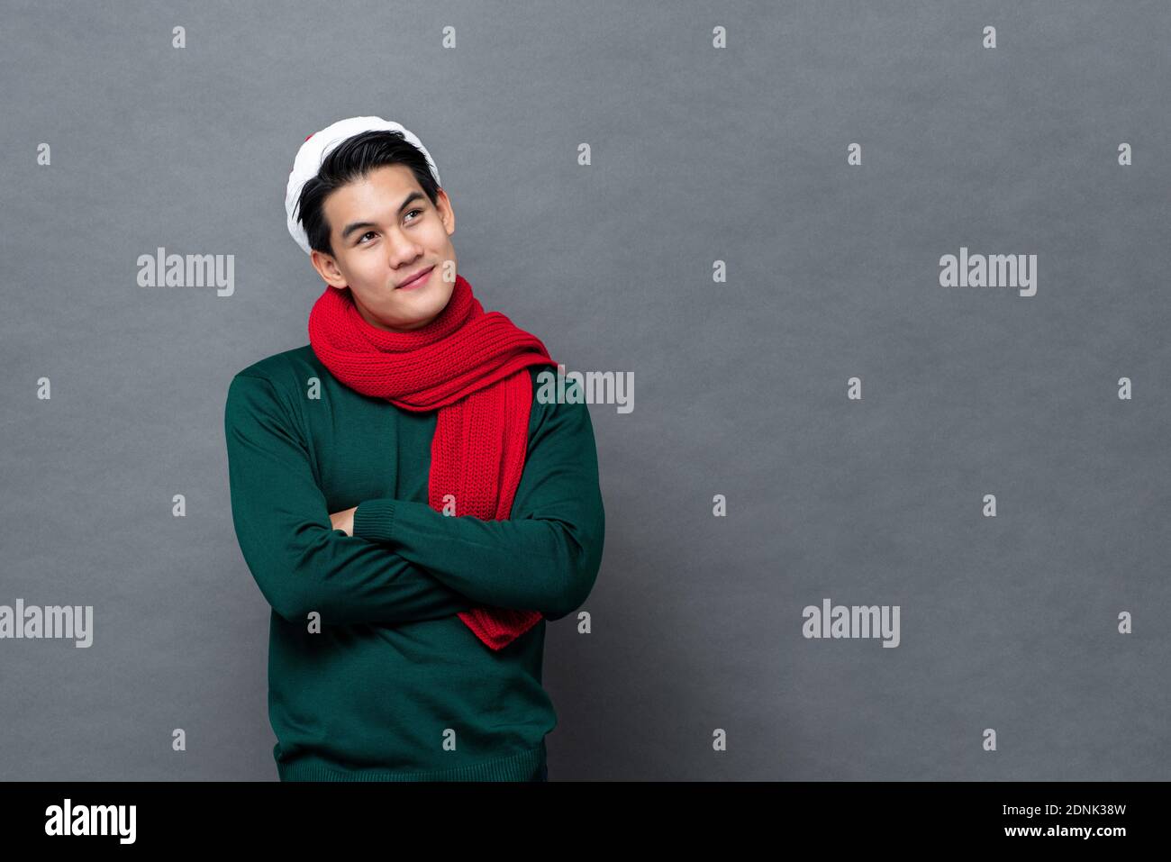 Junge schöne asiatische Mann in grün Weihnachten Pullover lächelnd und Blick nach oben auf den Kopierraum neben der Seite mit verschränkten Armen isoliert Auf grauem Hintergrund Stockfoto