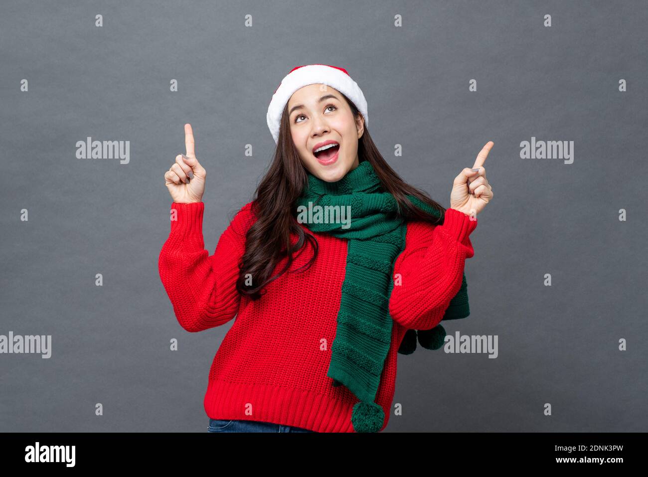 Glückliche asiatische Frau in rot und grün Weihnachten Kleidung suchen Und die Hände hoch in grau isoliert Studio Hintergrund Stockfoto