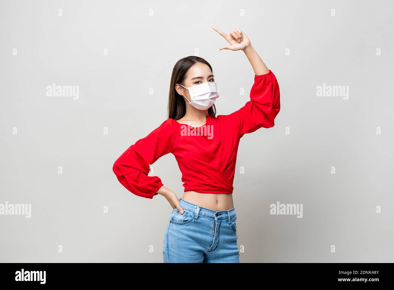 Junge asiatische Frau trägt medizinische Maske und poiting Hand nach oben Auf hellgrauem Studiohintergrund Stockfoto