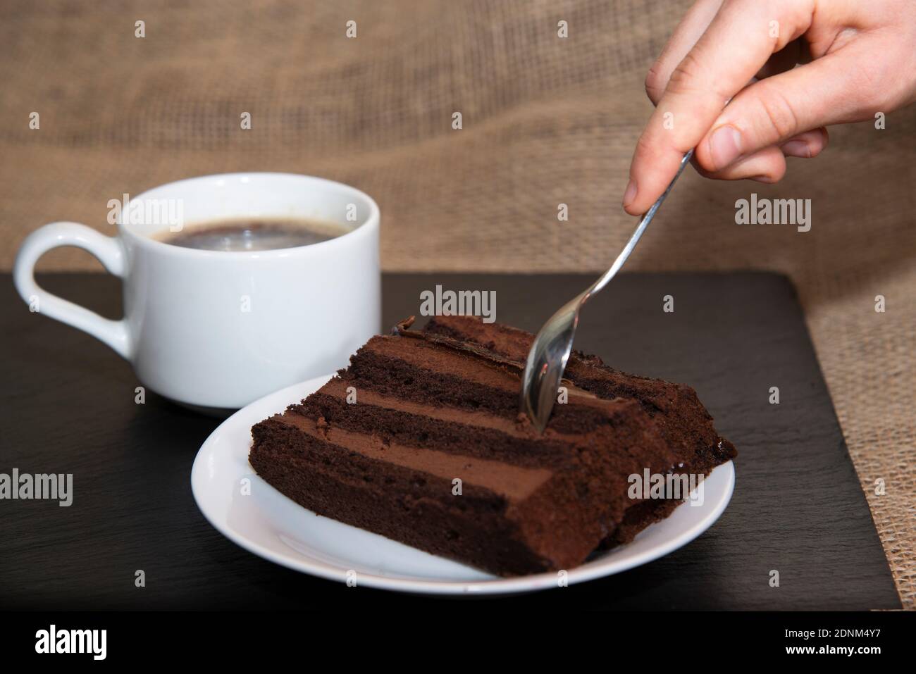 Eine Frauenhand hält einen Dessert-Metalllöffel und nimmt ein Stück Schokoladenkuchen neben einer Tasse Kaffee. Das Konzept der süßen Kaffeepause mit Kuchen Stockfoto