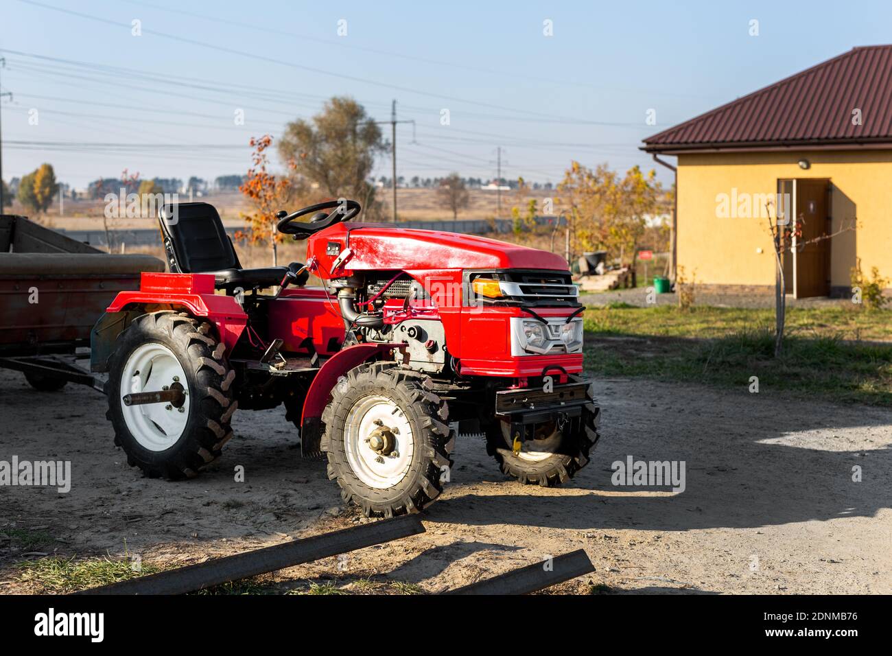 Kleiner Mini-roter moderner orangefarbener neuer Traktor mit Anhänger, der in der Nähe des Hangargebäudes auf dem Land der Farm bei Sonnenuntergang oder Sonnenaufgang steht. Kleine landwirtschaftliche Betriebe Stockfoto