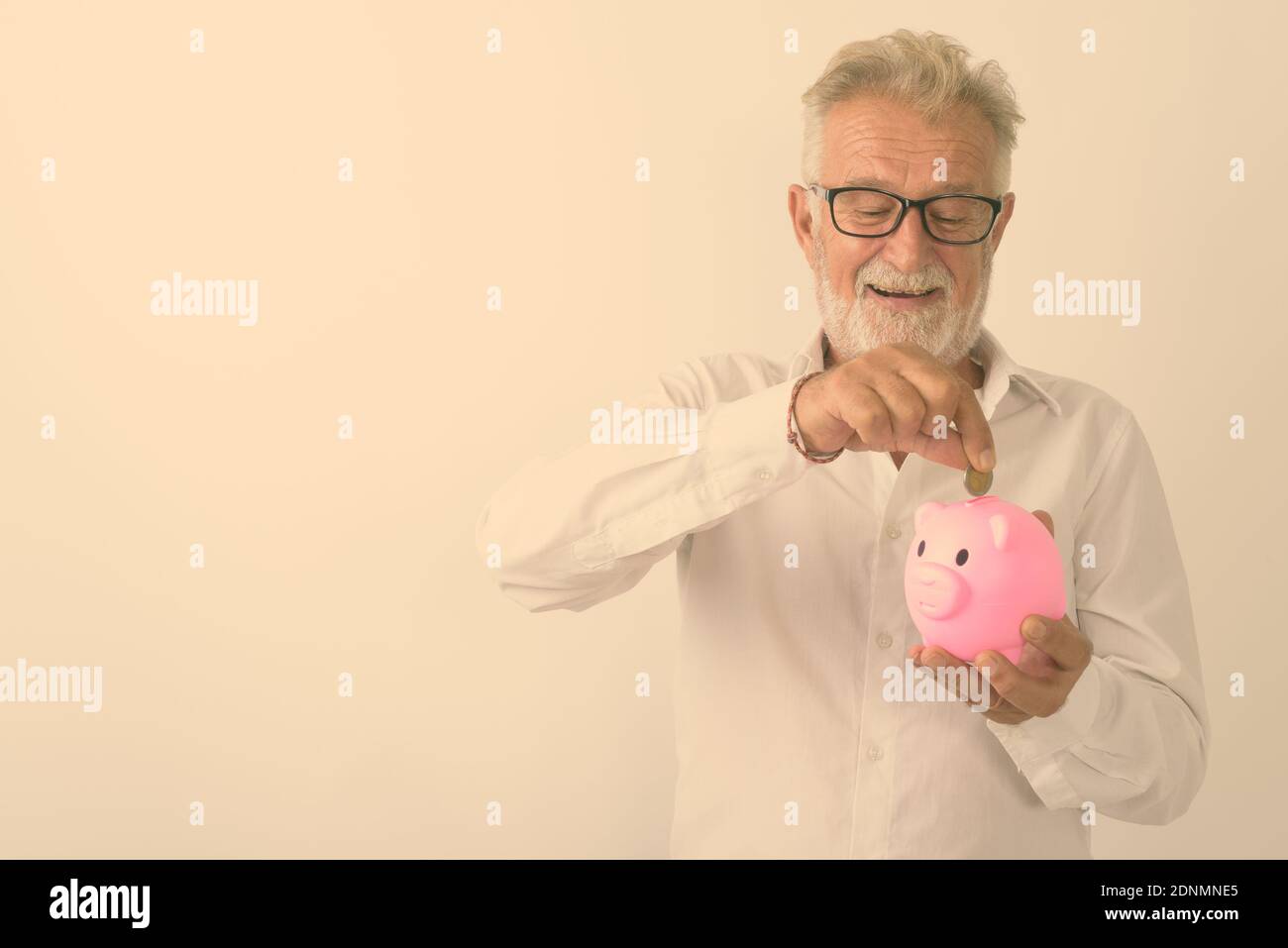 Studio shot von Happy älterer bärtiger Mann lächelnd, während sie und die Münze in Sparschwein mit Brille vor weißem Hintergrund Stockfoto