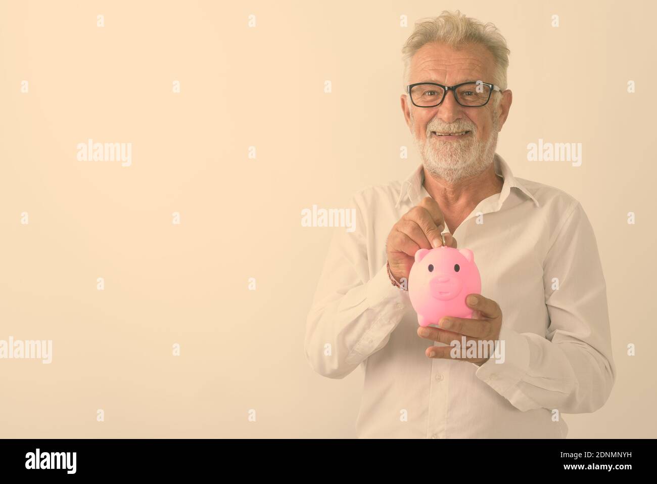 Studio shot von Happy älterer bärtiger Mann lächelnd, während sie Münze in Sparschwein mit Brille vor weißem Hintergrund Stockfoto