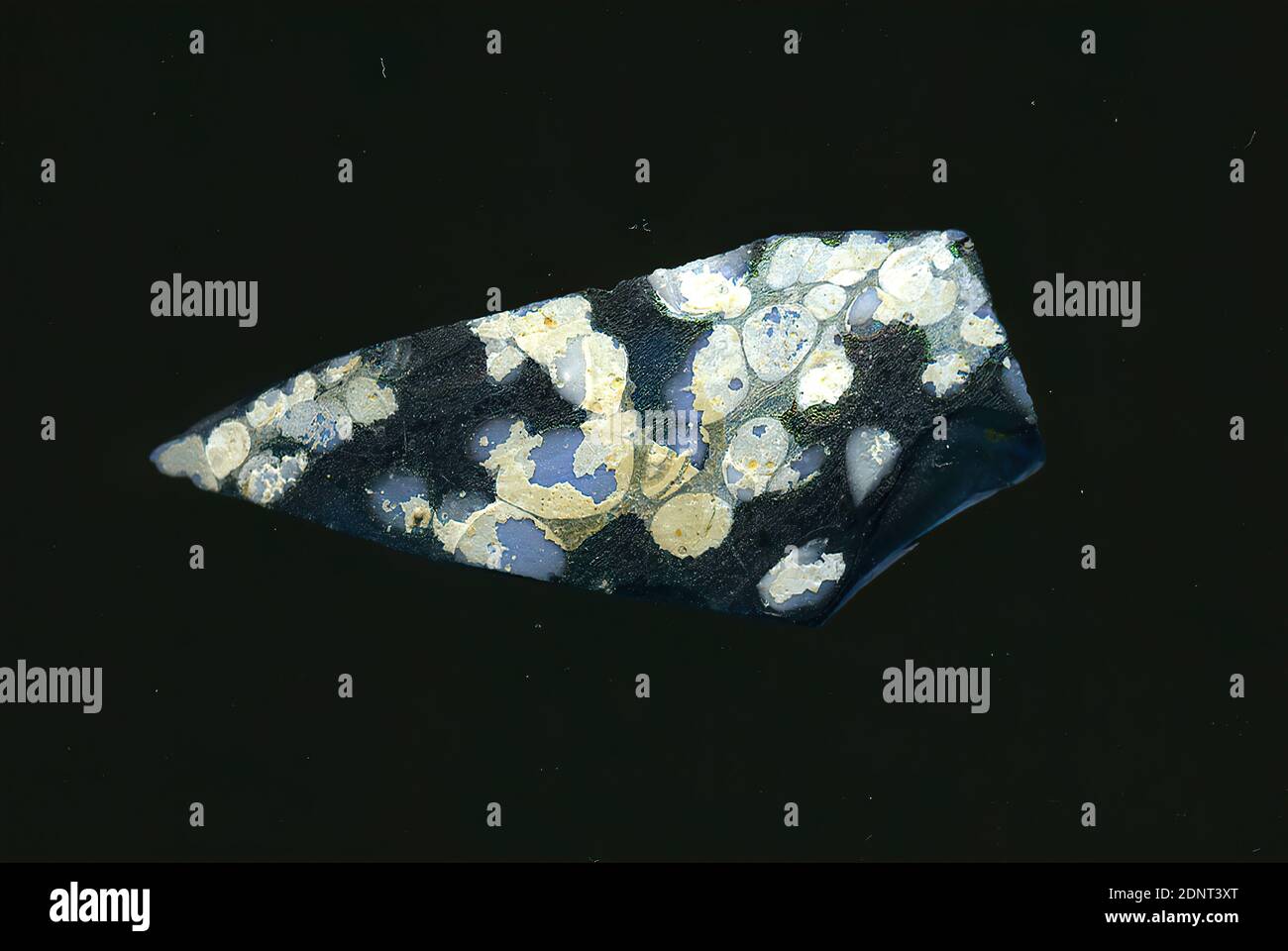 Fragment eines Gefäßes, Glas, gepresst, geschnitten, Gesamt: Höhe: 4.00 cm; Breite: 1.70 cm, frühe Kaiserzeit, das kleine Fragment des durchsichtigen blauen Glases ist unregelmäßig mit hellblauen Punkten durchsetzt. An diesen Stellen hat sich weiße Korrosion oder Sinterbildung gebildet. Für die Herstellung wurden verschiedenfarbige Gläser zu Rohren und Stäben geformt. Diese wurden zusammengedrückt und erhitzt, was zu einer langen Stange führte. Das in Scheiben geschnittene Glas hatte verschiedene amorphe oder florale Muster. Mehrere dieser Platten wurden dann zusammengefaltet, wieder erhitzt und in die Behälterform gepresst. Wegen der Anlage Stockfoto