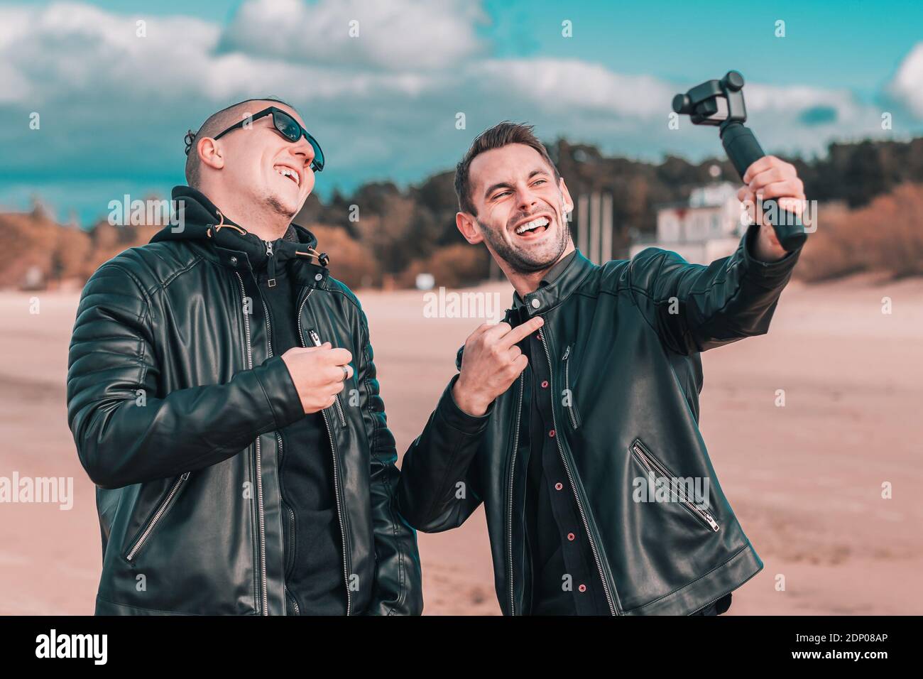 Zwei gutaussehende lächelnde Freunde machen Selfie mit Action-Kamera mit Gimbal Stabilisator am Strand. Jugendliche Männer in schwarzen Kleidern Spaß haben, indem Sie Fotos Stockfoto