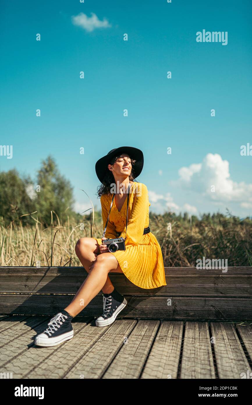 Junge Frau trägt einen schwarzen Hut und gelben Kleid mit einer analogen Kamera sitzen auf Holzsteg Stockfoto