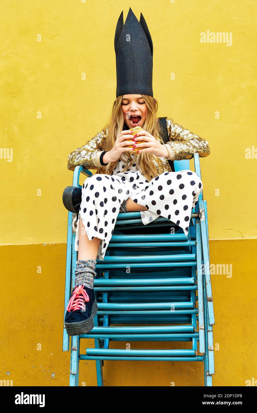Portrait von Mädchen mit schwarzen Krone sitzen auf Stapel Stühle essen Hamburger Stockfoto