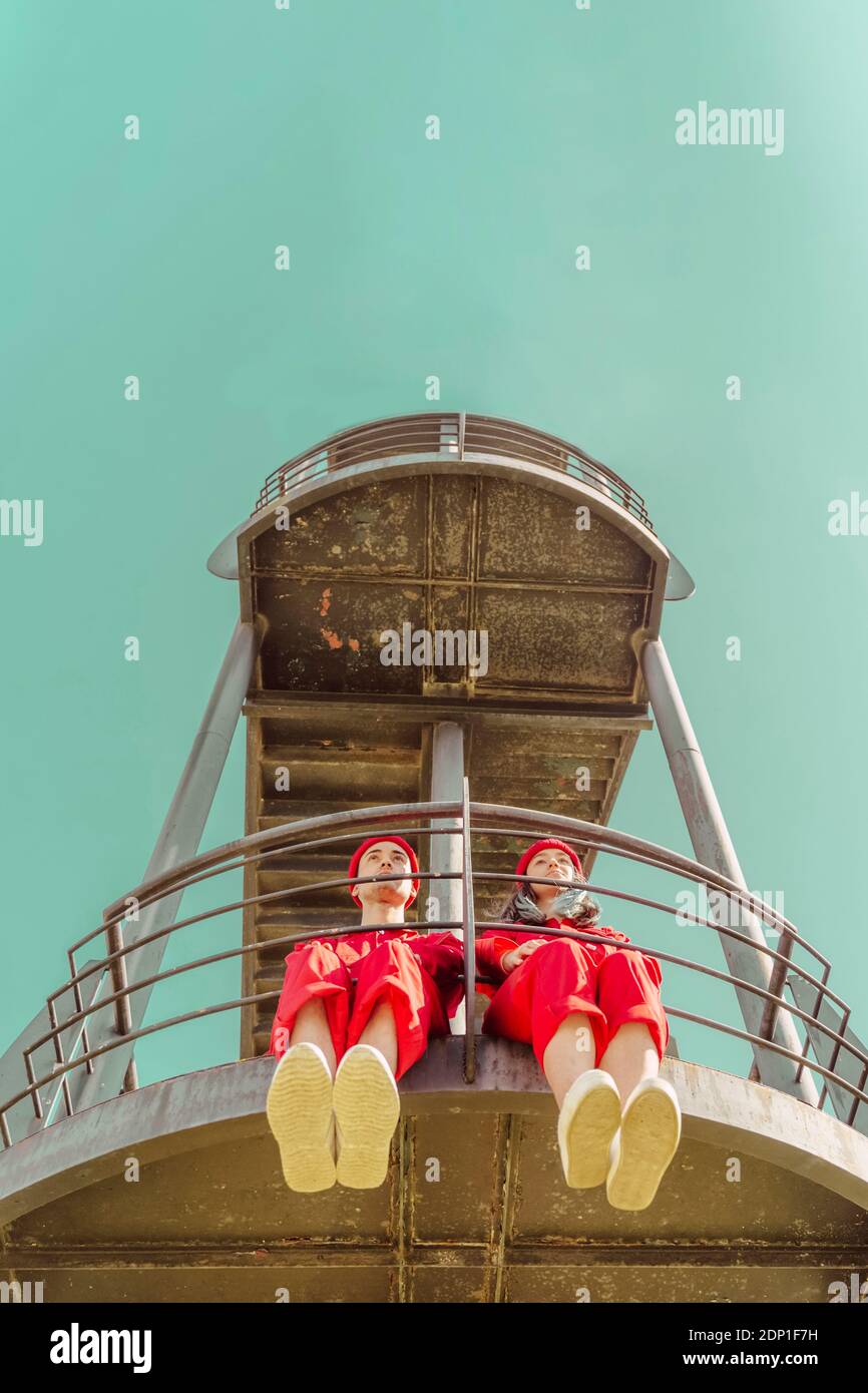 Junges Paar mit roten Overalls und Hüten, die nebeneinander sitzen Seite auf Plattform mit Blick auf die Entfernung Stockfoto