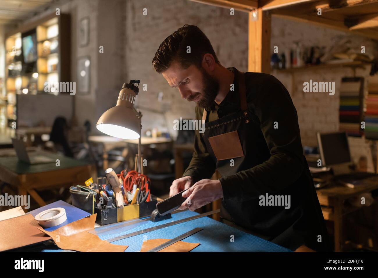 Bärtiger männlicher Handwerker, der während der Arbeit Werkzeuge über einem beleuchteten Tisch zubereitete Mit Leder im Heimstudio Stockfoto