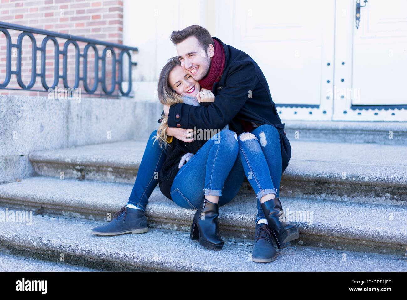 Porträt eines lächelnden Paares in Winterkleidung umarmt sitzend auf Einige Treppen auf einem städtischen Park Winterszene Stockfoto