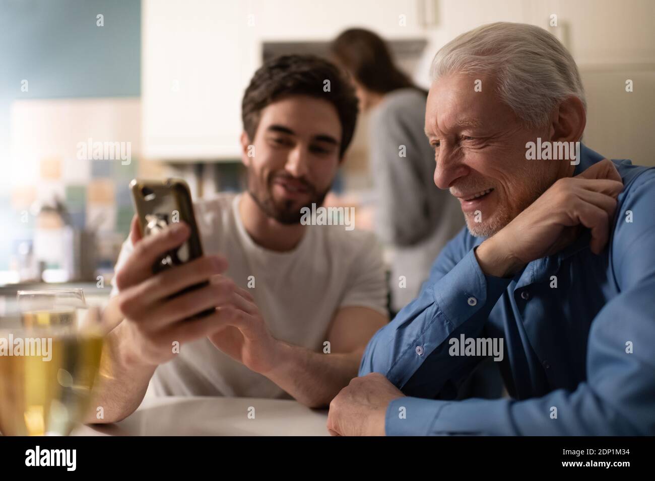 Älterer Vater lächelt und Fotos auf dem Smartphone von glücklich Junger Mann während des Familienessen in der Küche Stockfoto