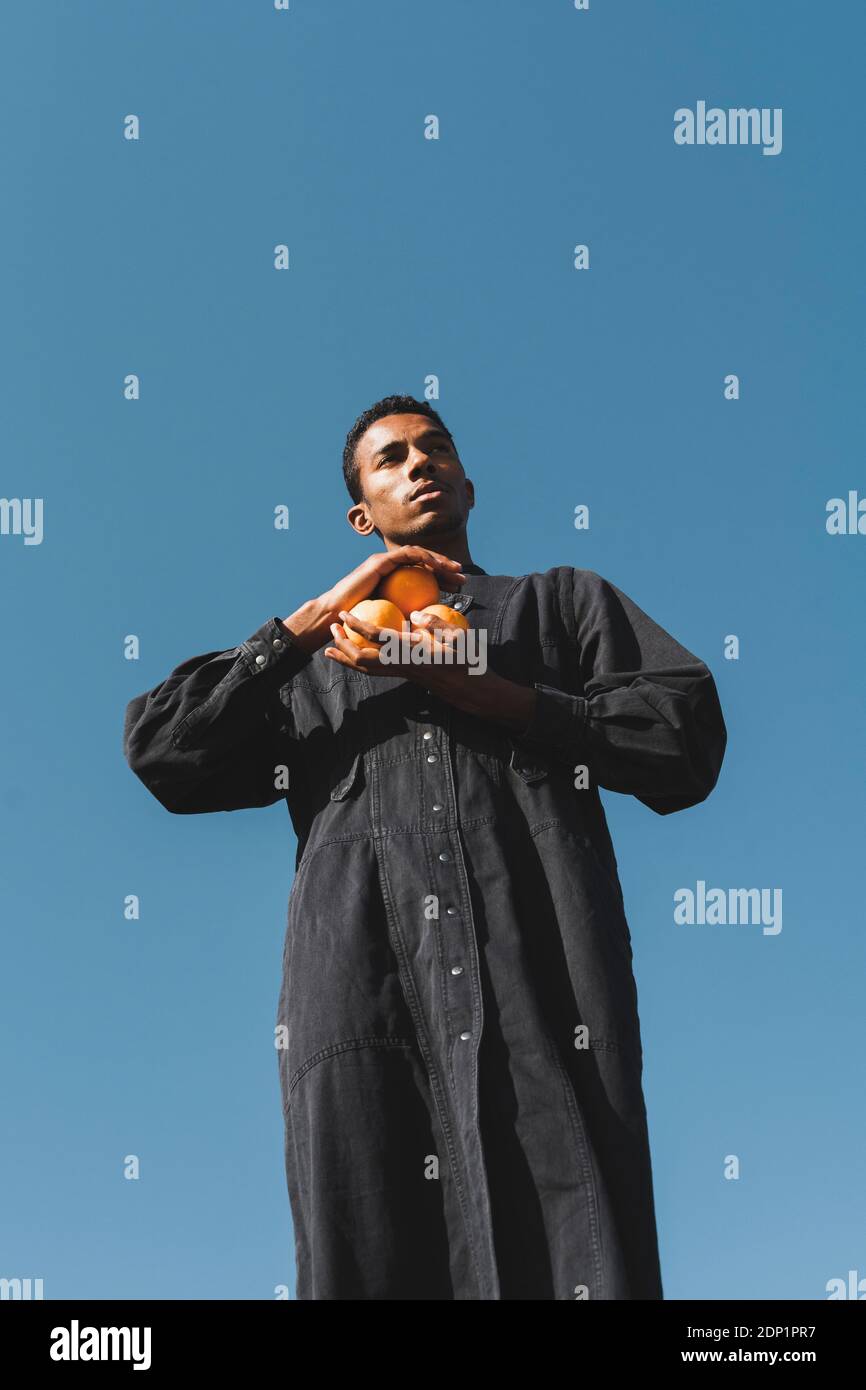 Junger Mann mit schwarzem Kaftan unter blauem Himmel, der Orangen hält Stockfoto