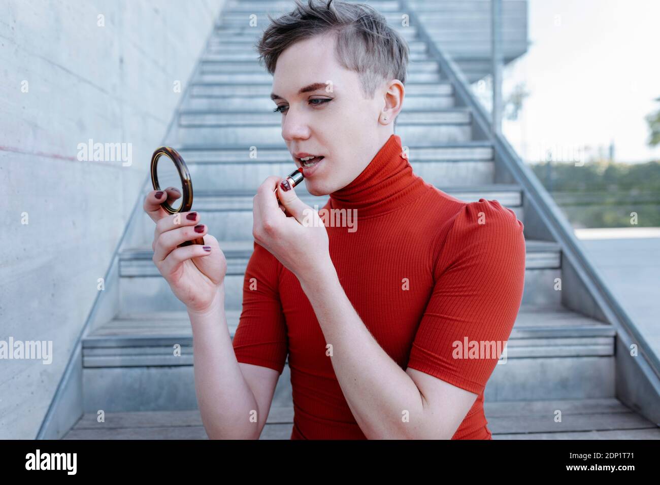 Nichtbinäre Person in einem roten Kleid Malerei ihre Lippen und Blick in einen Spiegel in