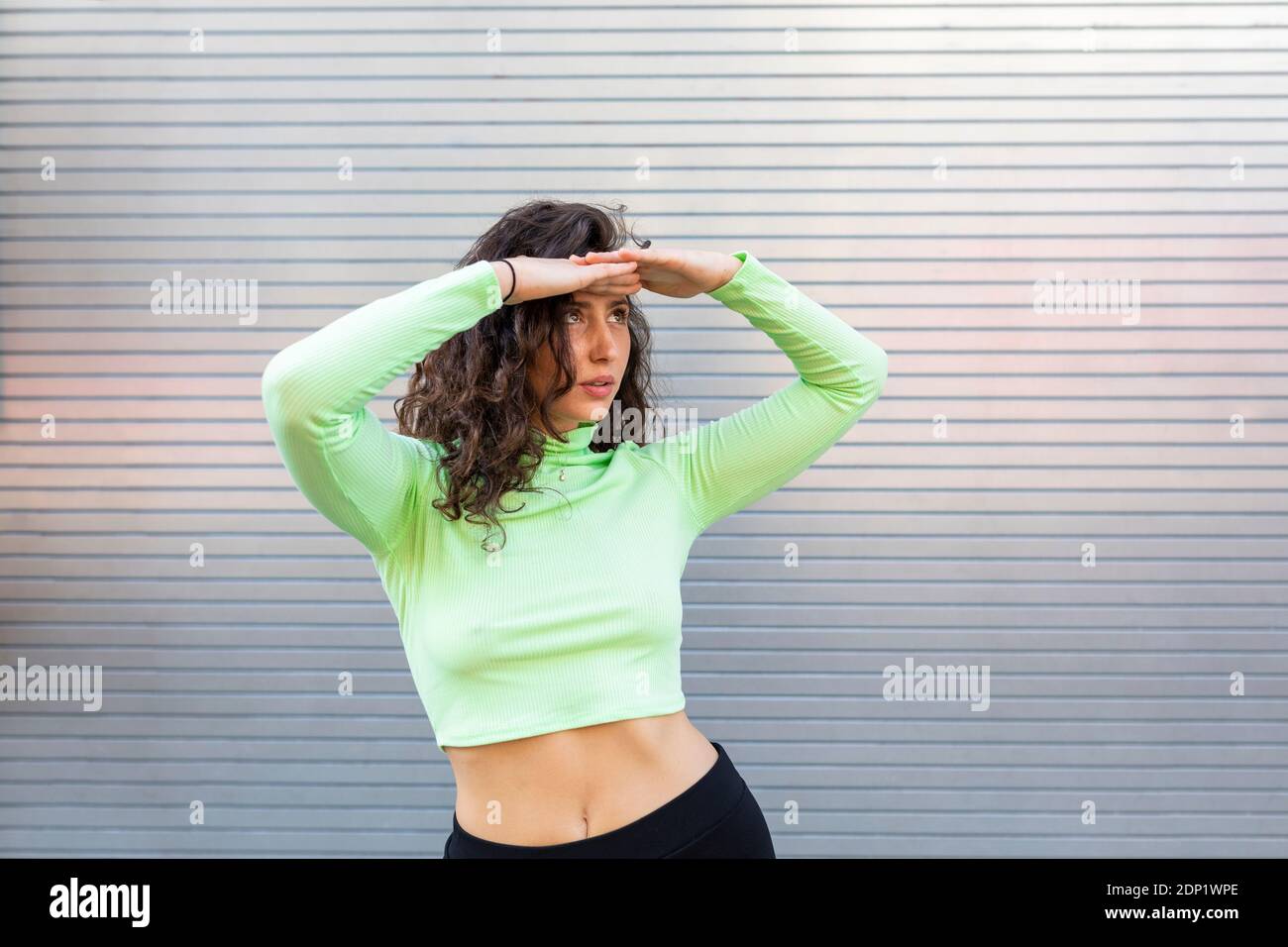 Junge Frau, die Augen abschirmt, während sie sich gegen die graue Wand posiert Stadt Stockfoto