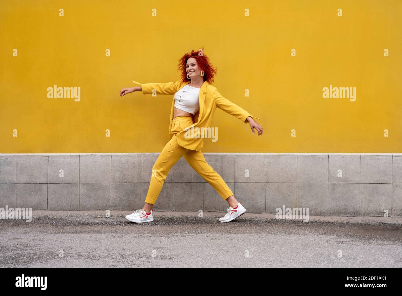 Energiegeladene junge Frau in gelbem Anzug läuft und springt hinein Vorderseite der gelben Wand Stockfoto