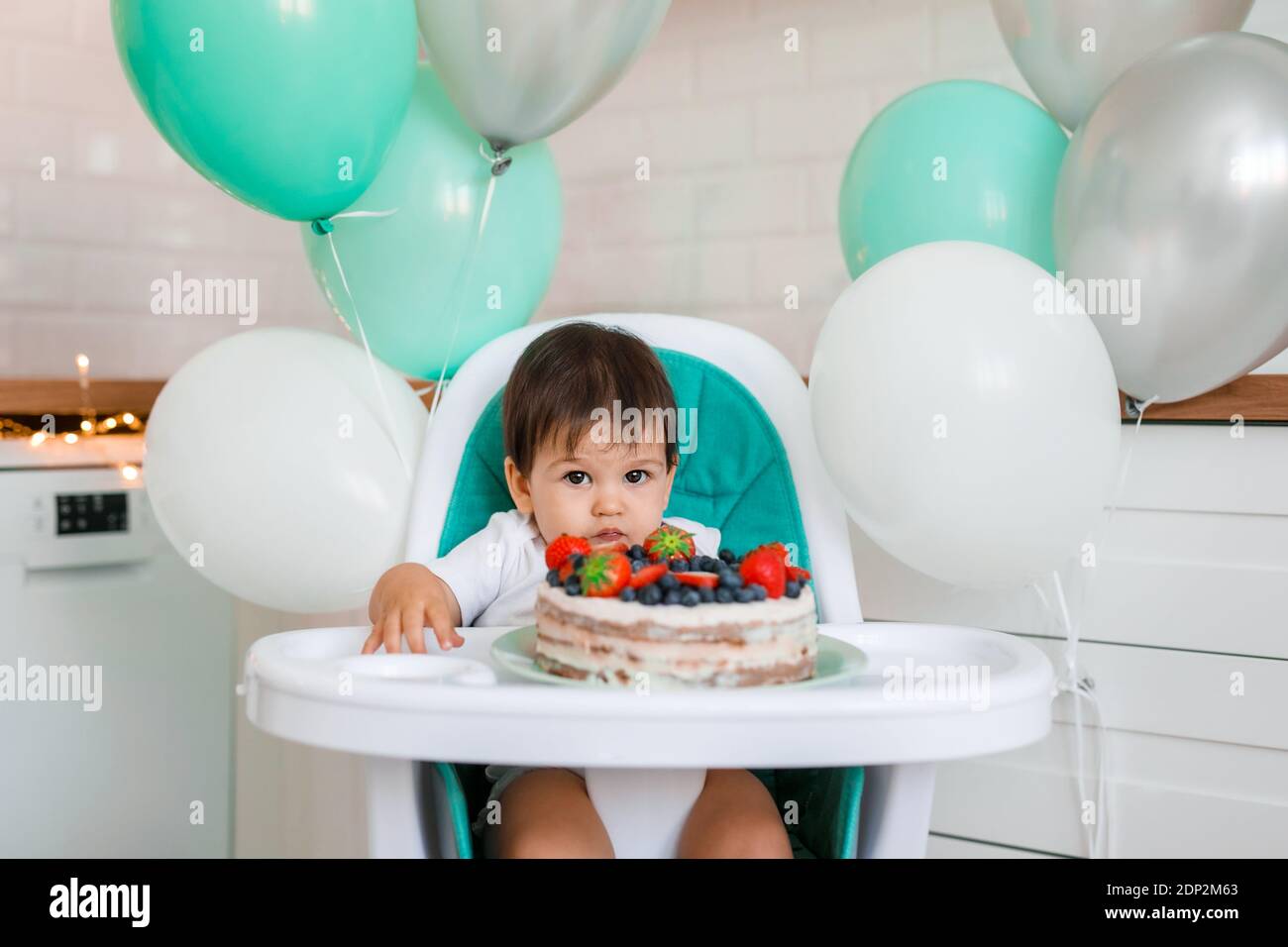 Kleiner Junge, der in einem Hochstuhl in der weißen Küche sitzt Und Verkostung ersten Jahr Kuchen mit Früchten auf Hintergrund mit Ballons Stockfoto