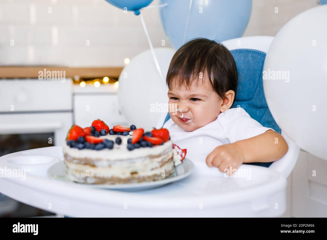 Kleiner Junge, der in einem Hochstuhl in der weißen Küche sitzt Und Verkostung ersten Jahr Kuchen mit Früchten auf Hintergrund mit Ballons Stockfoto