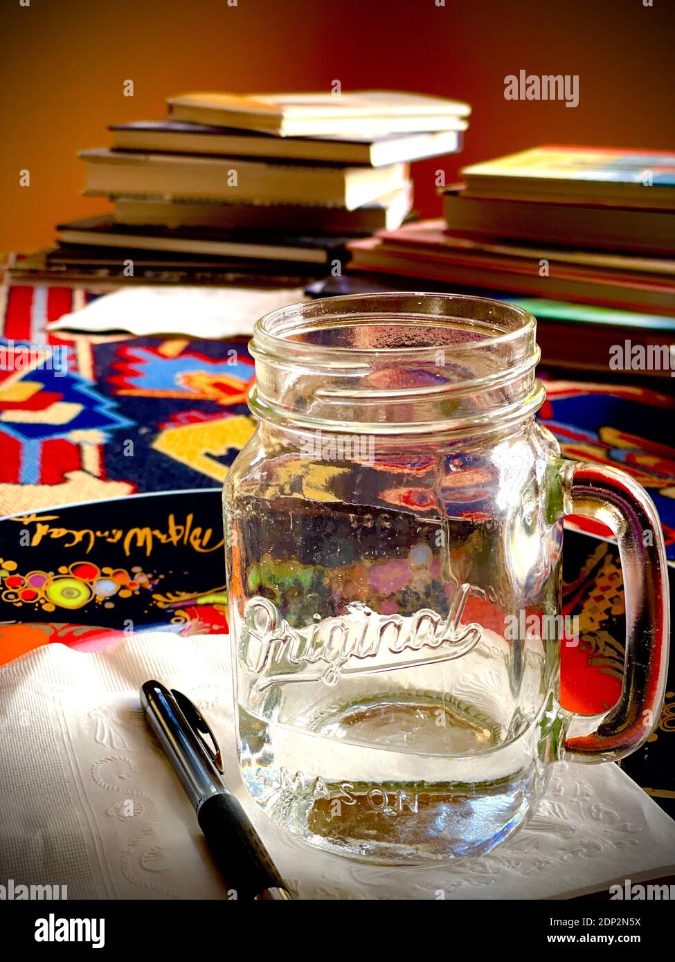 Original Mason Glas mit Griff auf buntem Material mit Stapel von Büchern im Hintergrund. Hintergrundbeleuchtetes, vertikales Farbfoto Stockfoto