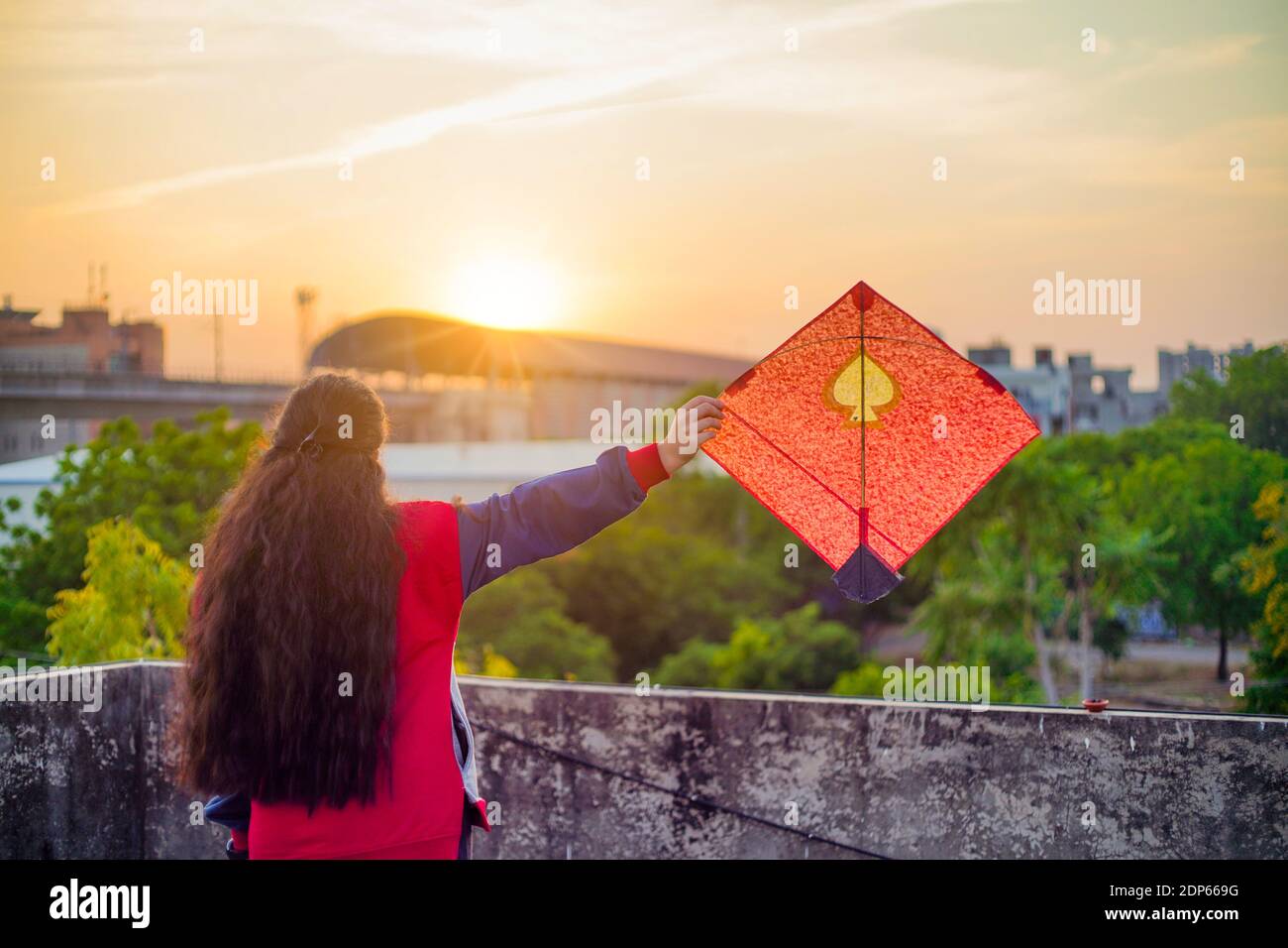 Junge Frau hält hoch bunte Papier und Holz Drachen gegen Ein verschwommener Hintergrund, der die Sonne auf dem indischen Drachenfest untergeht Von makar sankranti oder Stockfoto