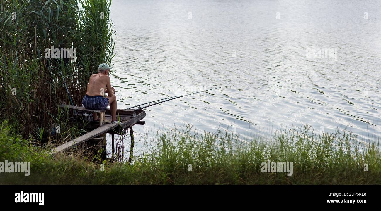 Ein männlicher Fischer sitzt auf einer hölzernen Brücke und Fische spinnen im Fluss mit Wasser in der Nähe des Schilf an einem Sommertag. Stockfoto