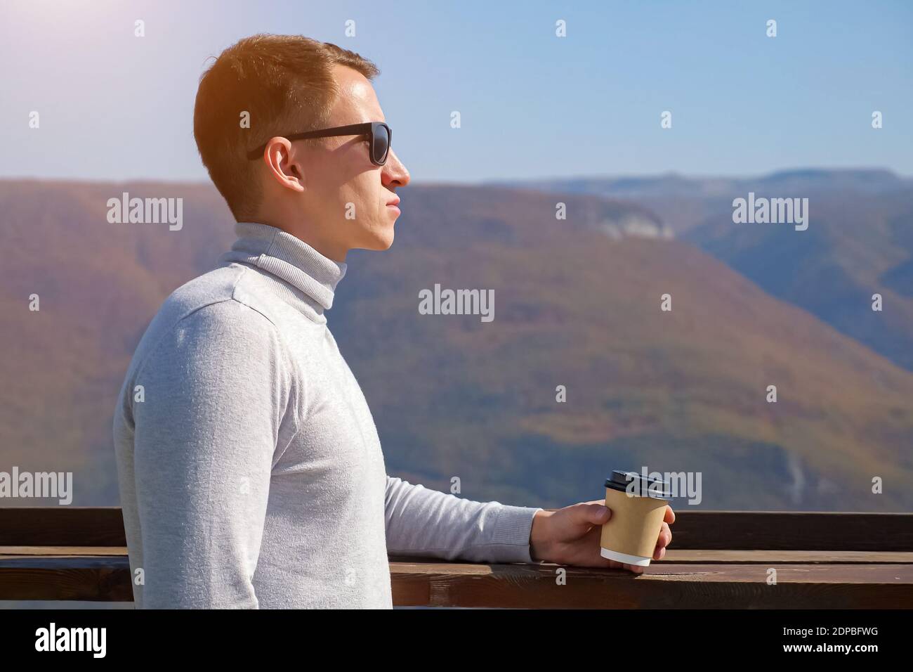 Junger Mann in einem grauen Rollkragen und Sonnenbrillen trinkt an einem klaren Tag, Sonnenlicht, ein Getränk aus einem Plastikglas auf einem Hintergrund von Bergen Stockfoto