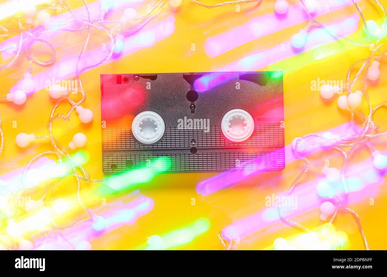 Retro vhs Videoband mit leuchtend leuchtenden LED Girlanden auf einem gelben Hintergrund. 80er Jahre, Lichteffekt. Aussicht von oben, minimalistisch Stockfoto