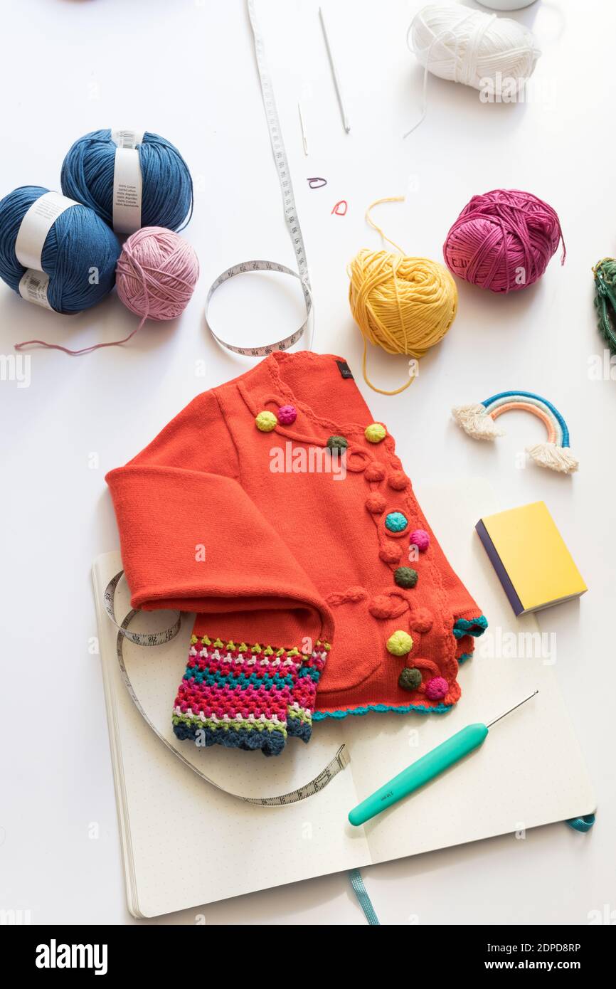 Eine bunte Strickjacke mit Häkelmuster auf einem weißen Tisch, umgeben von bunten Garnkugeln und einem gestrickten Regenbogen. Stockfoto