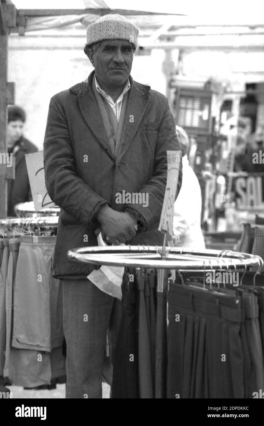 1970er Jahre, historisch, auf einem britischen Outdoor-Markt, ein männlicher Stallhalter aus dem Nahen Osten, der einen traditionellen ethnischen islamischen runden Hut trägt, an einem runden Stahlständer stand, der weibliche Kleidung verkauft, in England, Großbritannien. Stockfoto